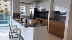 Una cocina moderna con armarios blancos, encimera de granito y electrodomésticos de acero inoxidable. Hay dos taburetes de bar y una pequeña isla con un frutero y un farol.