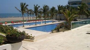 Un área de piscina con palmeras, una playa y un edificio con balcones.