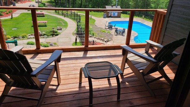 En træterrasse med to Adirondack-stole og et lille bord, med udsigt over en swimmingpool og et anlagt område med et picnicbord og bænke.