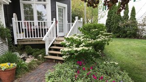 Une maison avec un porche à l’avant, un chemin en briques et un jardin où poussent différentes plantes et fleurs.