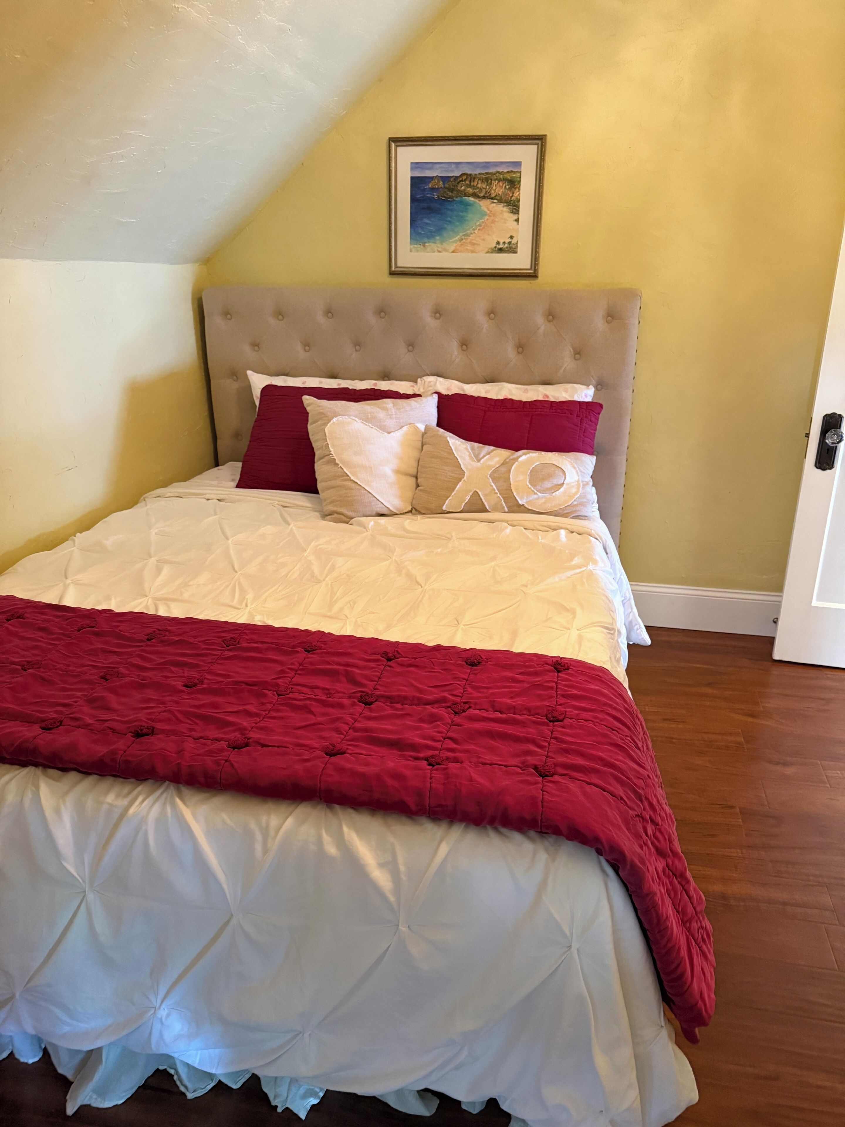 A bedroom with a tufted headboard, a bed with a white and red quilt, and a framed picture on the wall.