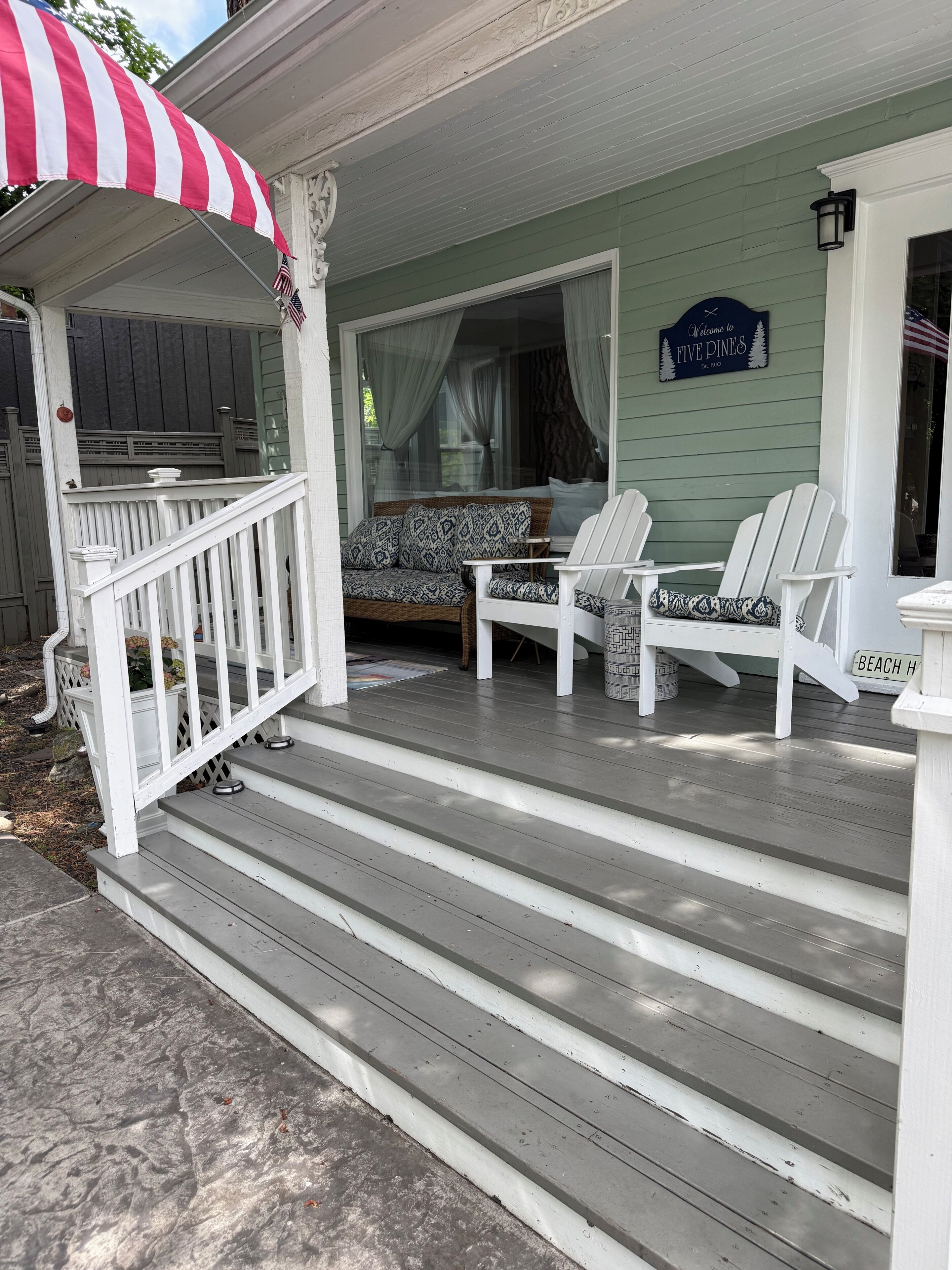 A porch with a white railing, a bench, two Adirondack chairs, and a sign that reads 'Welcome to Five Pines.'