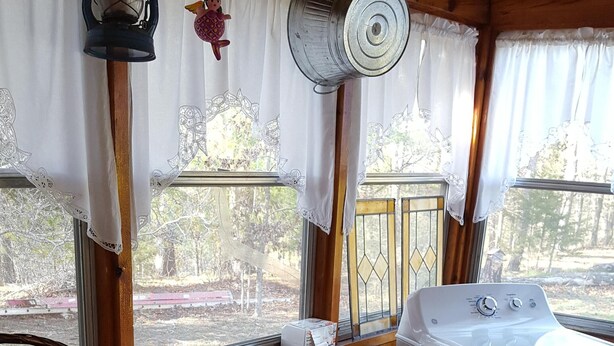 Interior de una cabaña con lavadora, una mesa con una cesta, un farol y una ventana con cortinas.