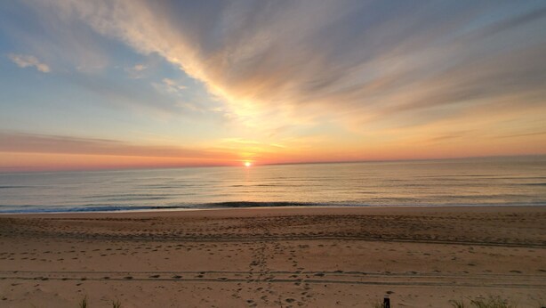 A beach with footprints leading to the water, a clear sky, and the sun setting over the horizon. - OCEANFRONT☆One level living w/ramp access to house & dune deck. Walk to dining!  (Kitty Hawk)