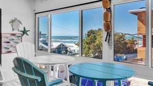 A room with large windows, a white chair, a blue table, and a view of the beach and ocean. - Sandy Cheeks & Barefeet, Gorgeous interior Gulf beach Place, pool, on beach side (Holmes Beach)