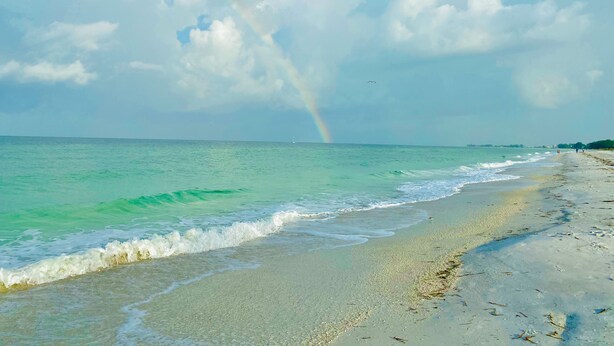A beach with clear turquoise waters, a visible rainbow, and a distant sailboat.