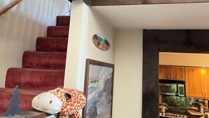 A staircase with a wooden handrail, a plush toy on the wall, a framed picture, and a kitchen area visible through an open door. - lake George Adirondack Chalet  (Lake George)