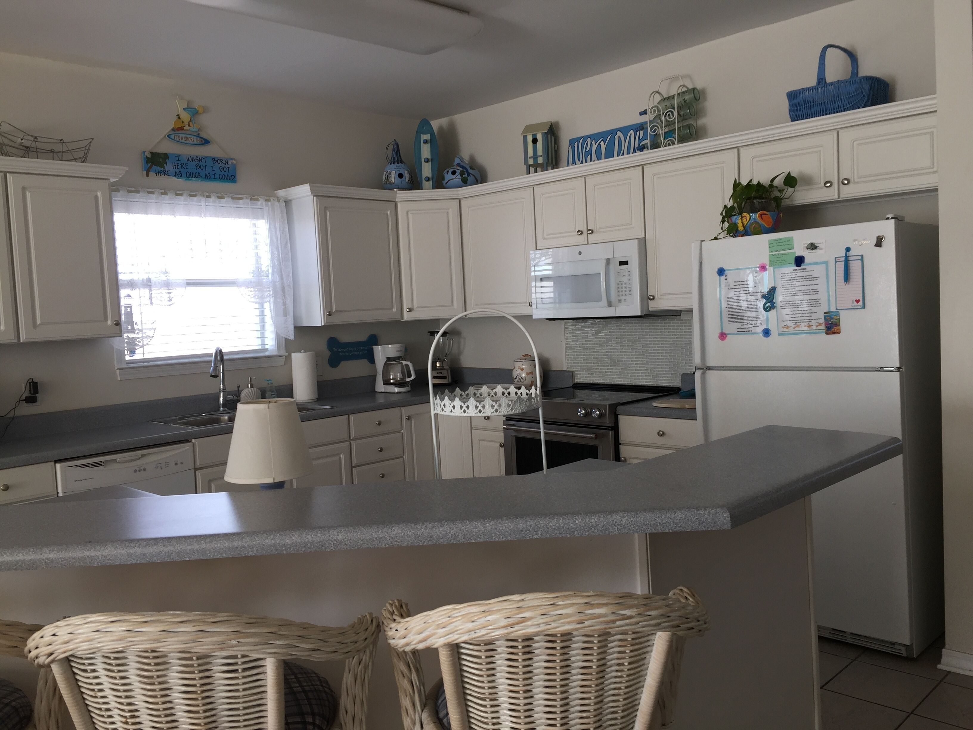 Una cucina moderna con mobili bianchi, un'isola centrale con due sedie in vimini, un forno a microonde e un frigorifero decorato con calamite e bigliettini.