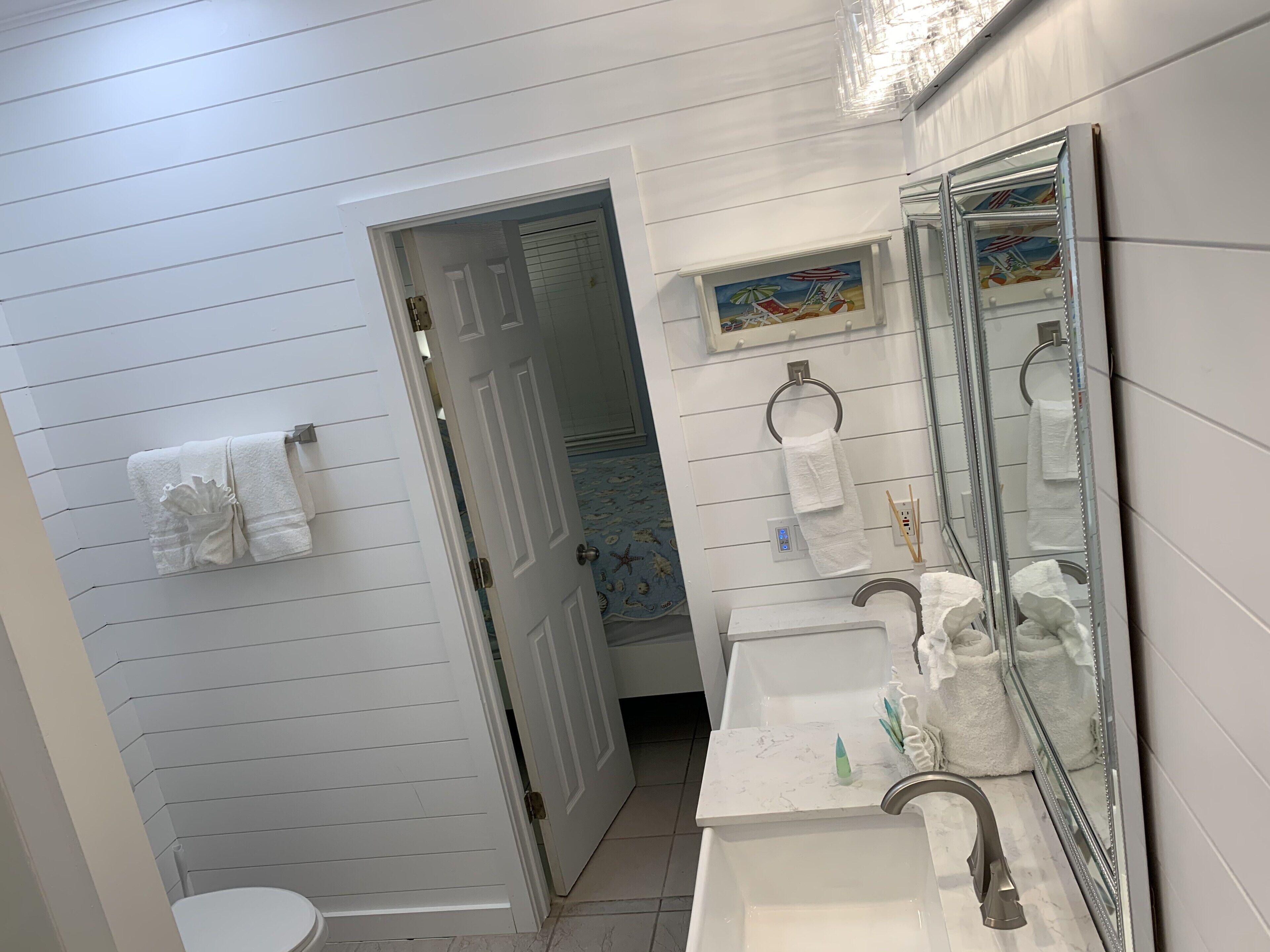 A bathroom with a toilet, sink, and mirror. There is a towel rack with towels and a painting above the mirror.