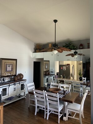 Uma sala de jantar espaçosa com uma mesa de madeira e cadeiras brancas, uma cozinha ao fundo e um ventilador de teto.