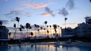 A swimming pool with lounge chairs, palm trees, and a sunset in the background. - Playa Turquesa, Beachfront, Ocean view, pool, 2 Bd. Luxury Condo, Wifi, Smart TV (Bavaro)