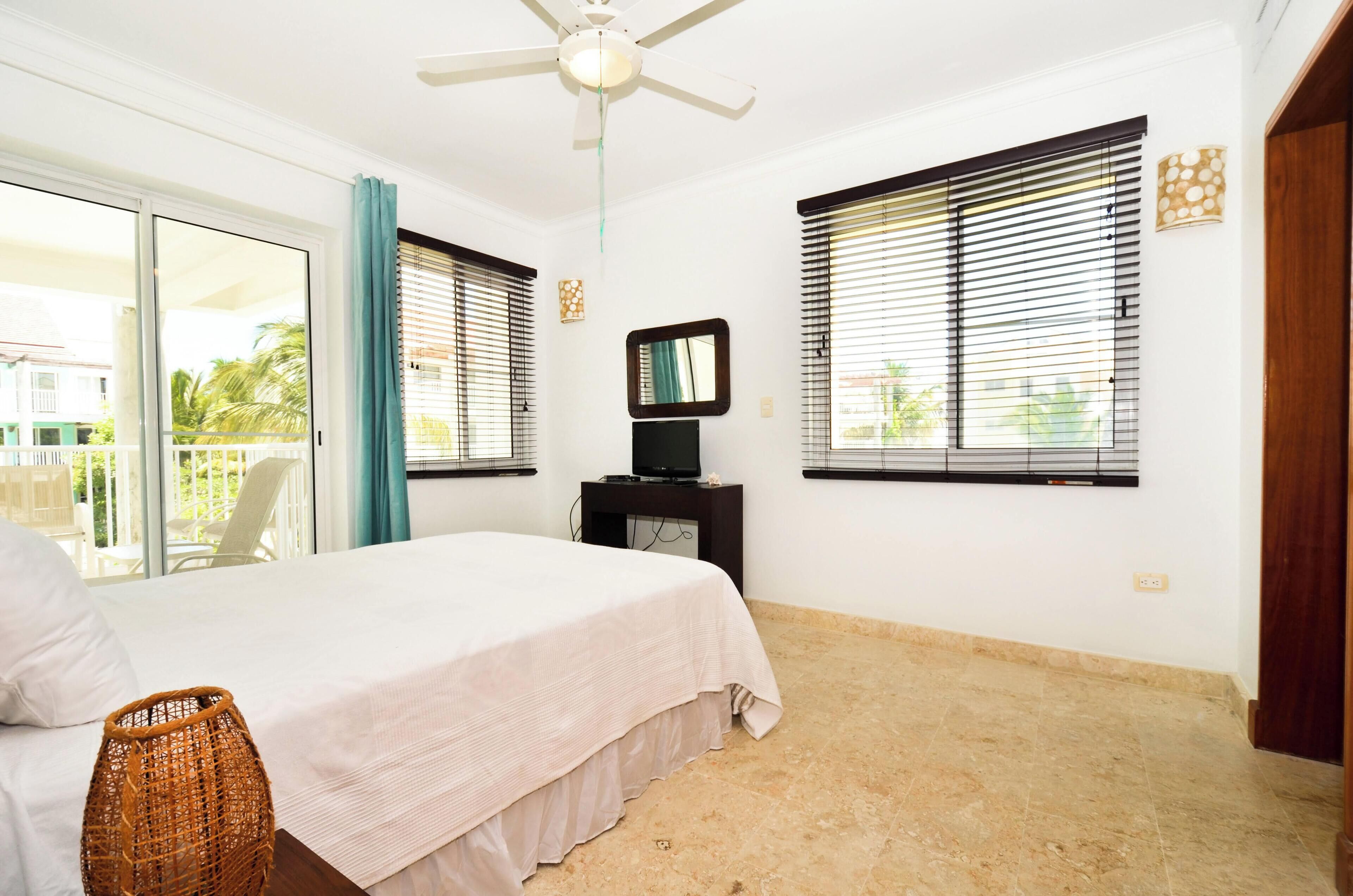 A bedroom with a bed, a ceiling fan, a TV on a stand, and large windows with blinds.