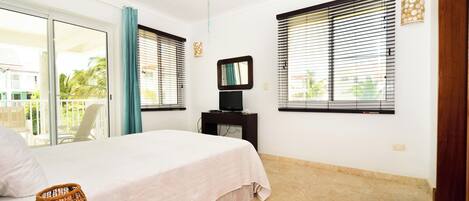 A bedroom with a bed, a ceiling fan, a TV on a stand, and large windows with blinds.