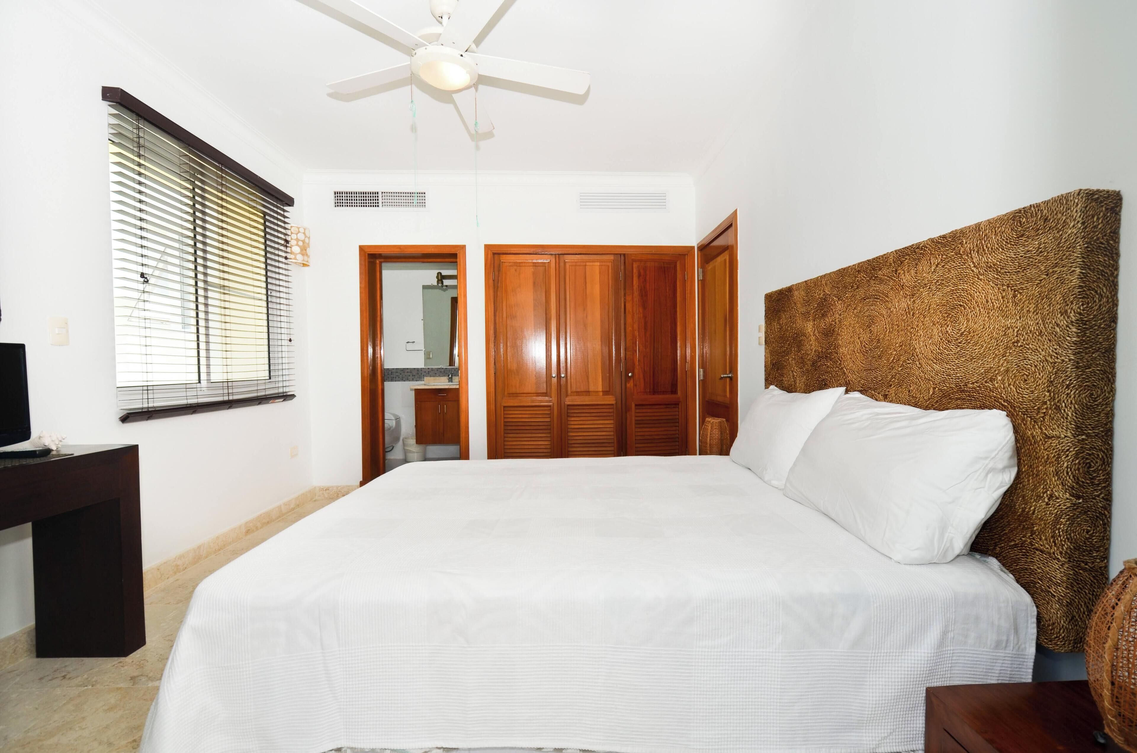 Une chambre à coucher avec un lit, une armoire en bois, un ventilateur de plafond et une fenêtre avec des stores.