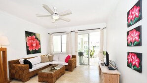 A living room with a sofa, coffee table, and a desk with a computer. There are large windows and a ceiling fan. - Playa Turquesa, Beachfront, Ocean view, pool, 2 Bd. Luxury Condo, Wifi, Smart TV (Bavaro)