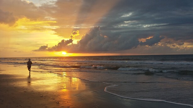 Cena de praia com pôr do sol, ondas e uma pessoa caminhando.