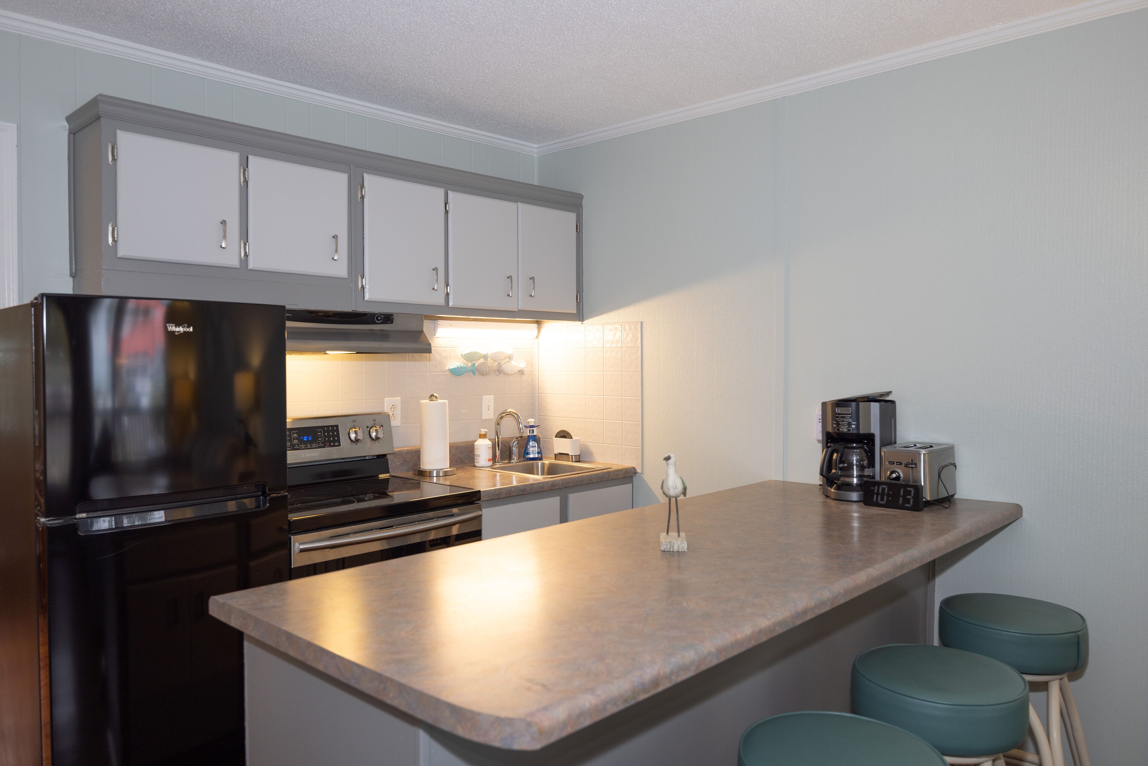A kitchen with a stainless steel refrigerator, a stove, a sink, and a coffee maker. There are three stools at the counter.