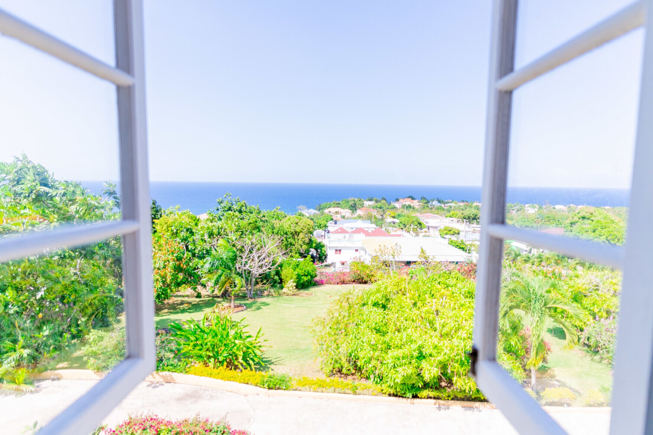 Une vue depuis une fenêtre donnant sur un jardin luxuriant, avec un bâtiment visible au loin et l’océan au-delà.