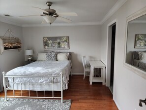 A bedroom with a bed, a desk, a chair, a ceiling fan, a mirror, and a painting on the wall.