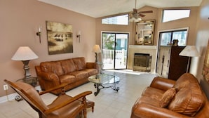 A living room with a brown leather sofa, a glass coffee table, and a wooden armchair. There is a fireplace, a ceiling fan, and a large window with a view of the outside. - beautiful Home 98 steps from the beach just off Thomas drive (Panama City)
