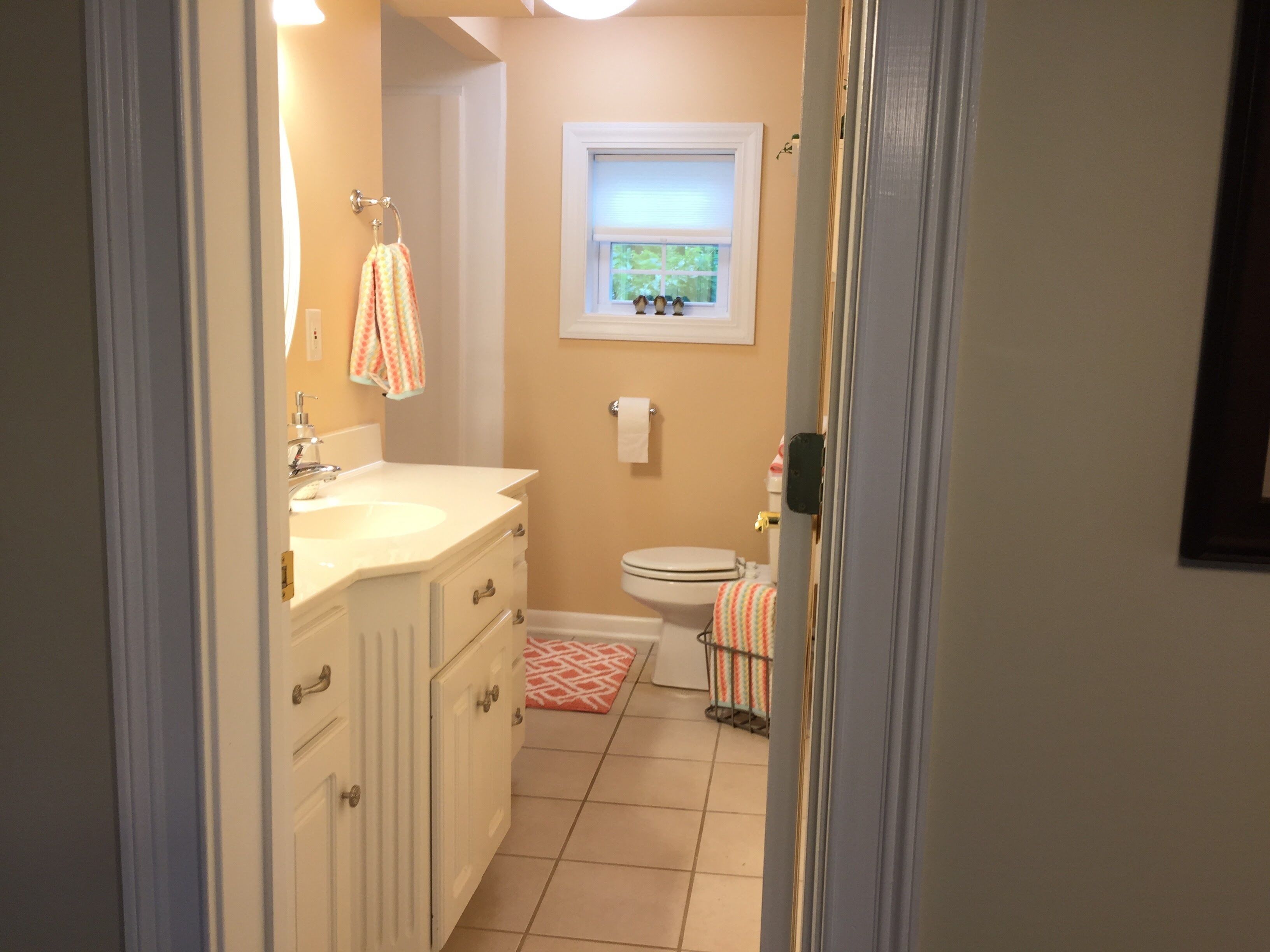 Baño con lavamanos, tocador y retrete blancos. Se ve una ventana con persianas y un perchero con una toalla colorida.