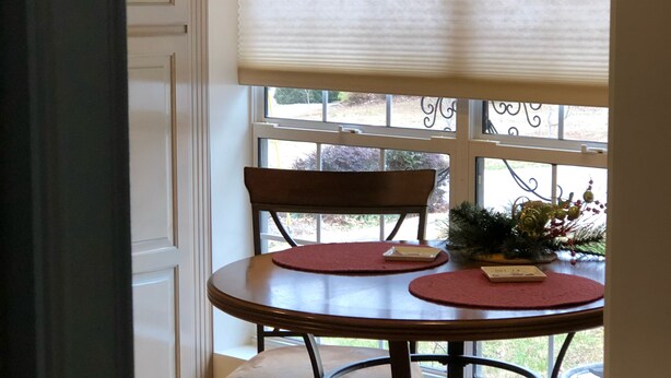 Un coin repas comprenant une table ronde et deux chaises, une fenêtre avec des stores et un élément décoratif mural placé au-dessus de la fenêtre.