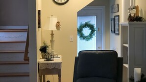 Un salon avec un fauteuil bleu foncé, une horloge murale blanche, une petite table avec une lampe et une guirlande accrochée à la porte.