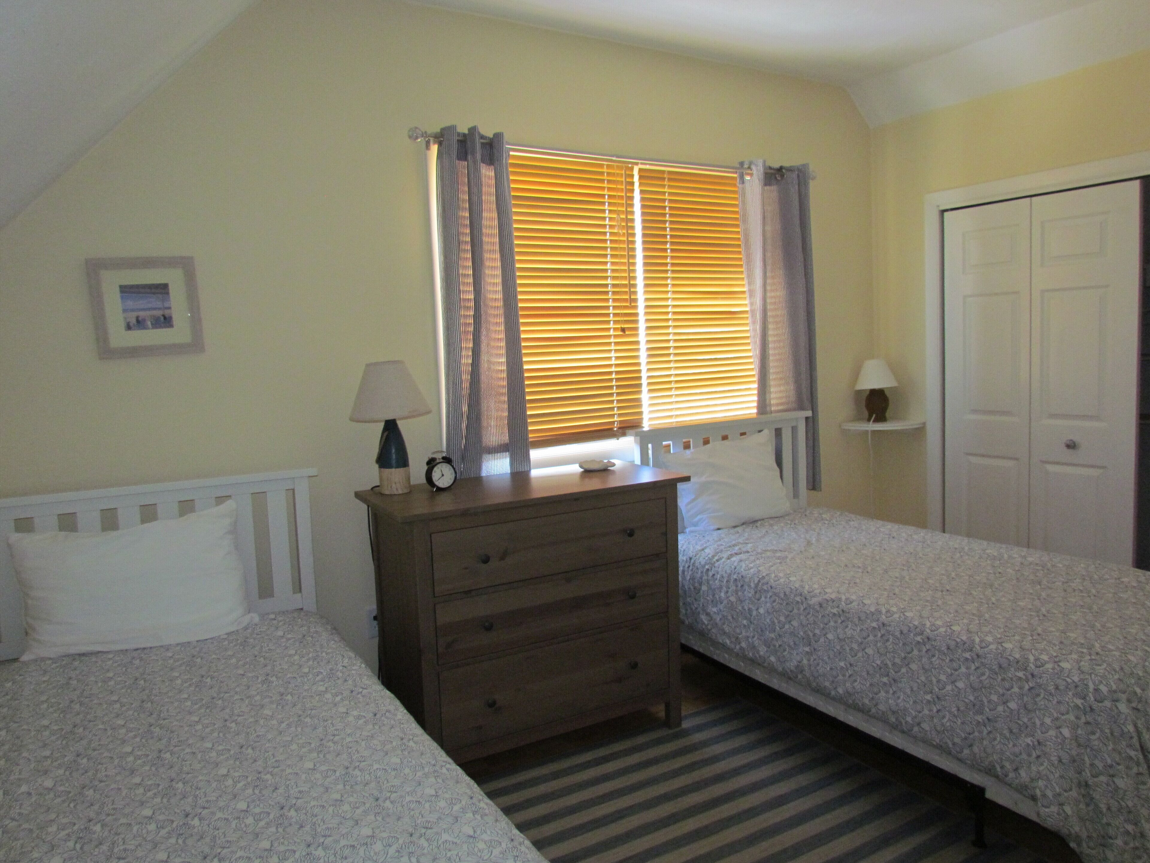 A bedroom with two single beds, a wooden dresser, a bedside table with a lamp, a window with blinds, and a door.