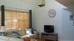 A living room with a beige sofa, a television on a stand, and a clock on the wall. - Vineyard Haven, Quiet Setting Just Minutes from Ferry and Bus (Vineyard Haven)