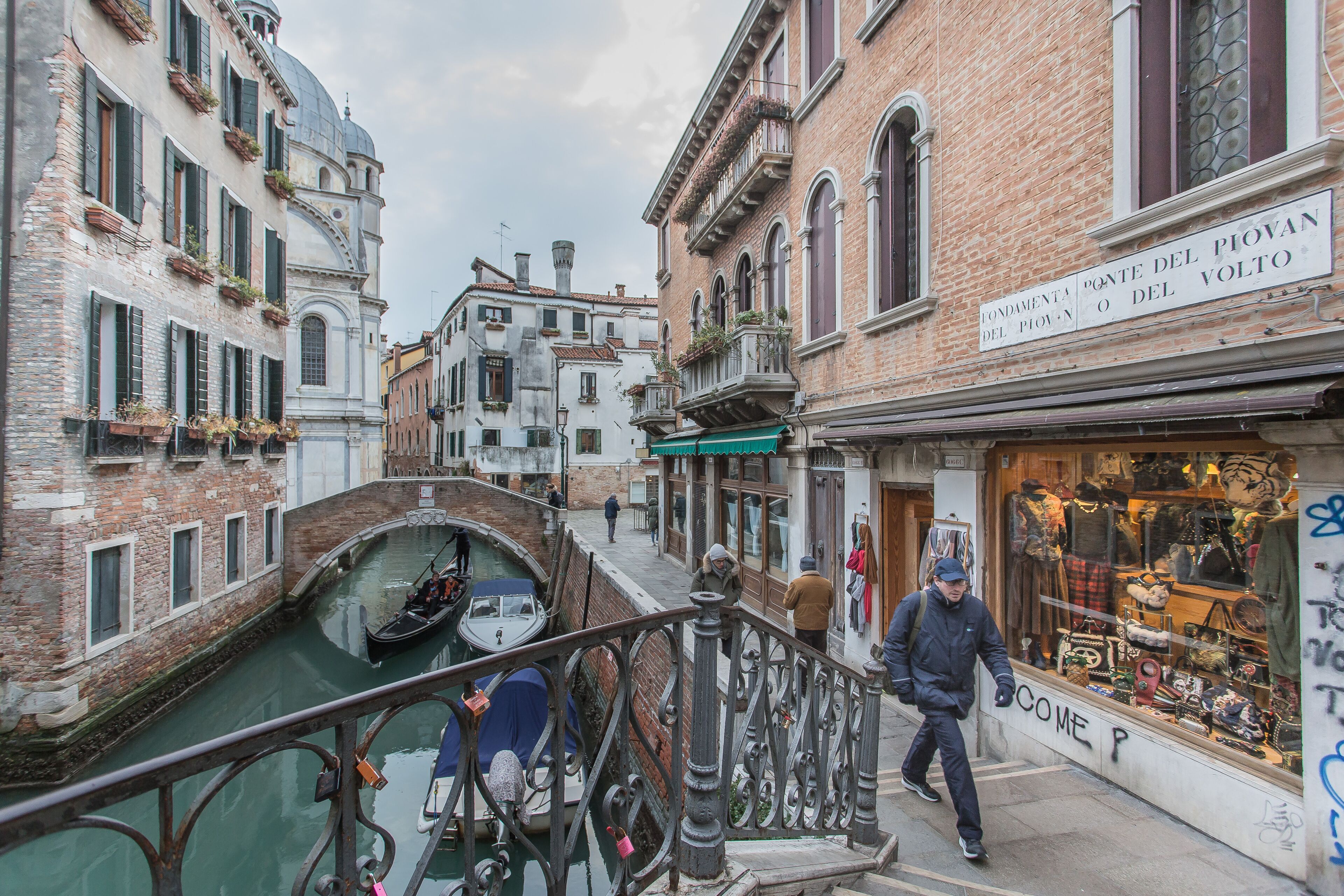 Un estrecho canal veneciano con una góndola, edificios con arquitectura tradicional veneciana y una tienda con un letrero en italiano.