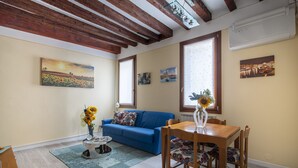 Uma sala de estar com um sofá azul, uma mesa de jantar de madeira e cadeiras, uma mesa de centro com um vaso de girassóis e duas janelas com cortinas.