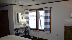 Een slaapkamer met een bed, een dressoir, een spiegel, een raam met gordijnen en uitzicht op een wateroppervlak.