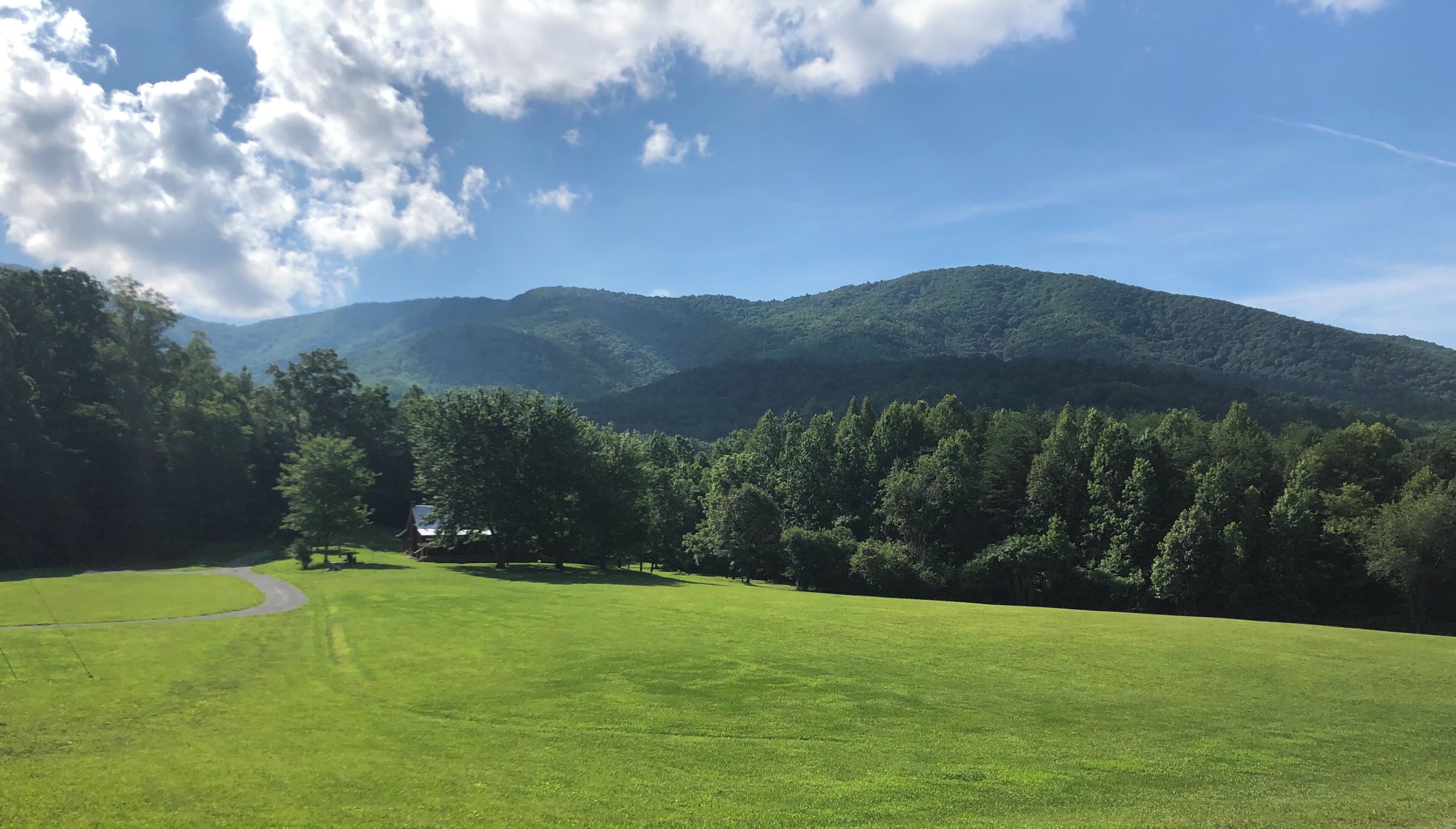 Un champ verdoyant avec un sentier menant à un bâtiment entouré d’arbres et de collines.