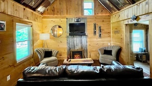 Ein gemütliches Blockhaus-Interieur mit Kamin, zwei Sesseln, einer Couch und einem Deckenventilator.