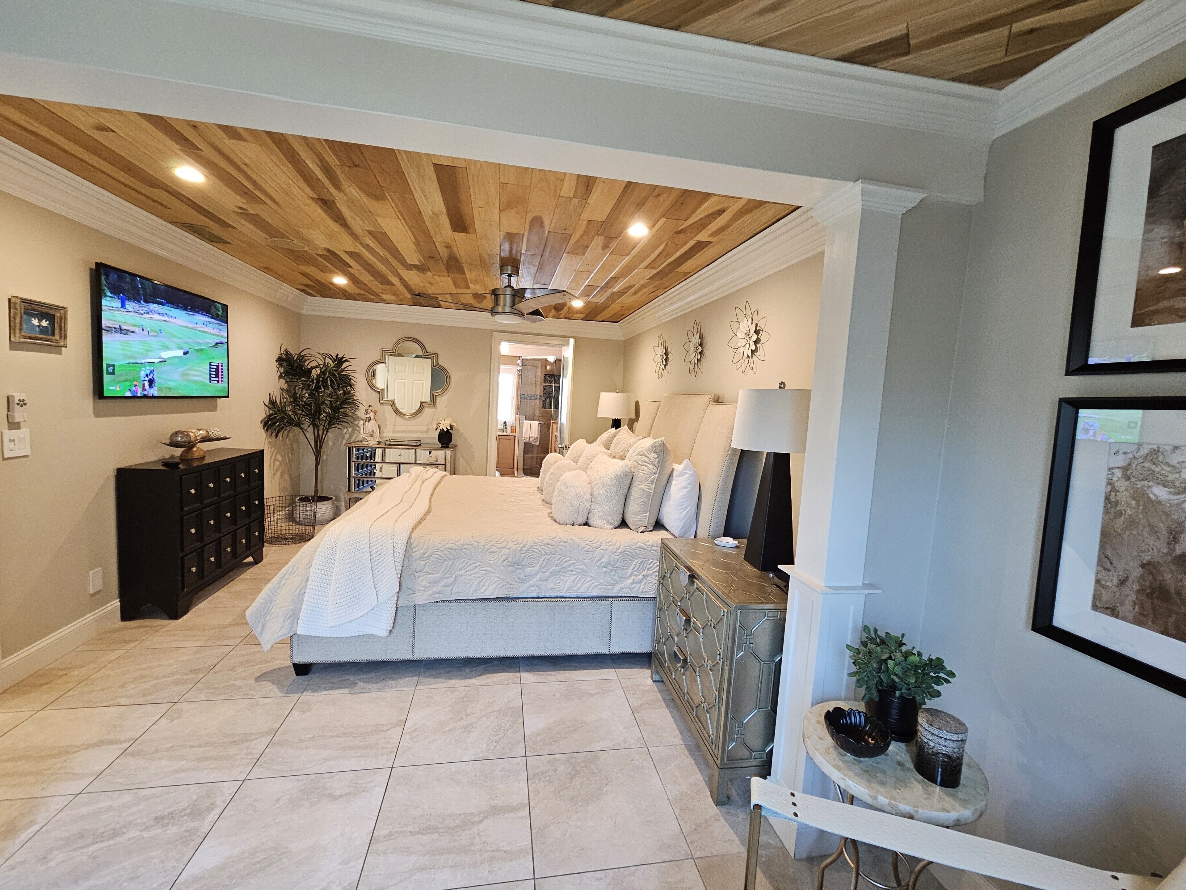 A bedroom with a wooden ceiling, a large bed, a TV on the wall, and a bathroom visible through an open door.