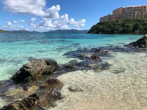 A coastal scene with clear turquoise waters, rocky shoreline, and a building with multiple floors in the background.