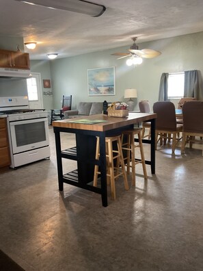 Een keuken met een centraal eiland, barstoelen, een eettafel met stoelen, een plafondventilator en een schilderij aan de muur.