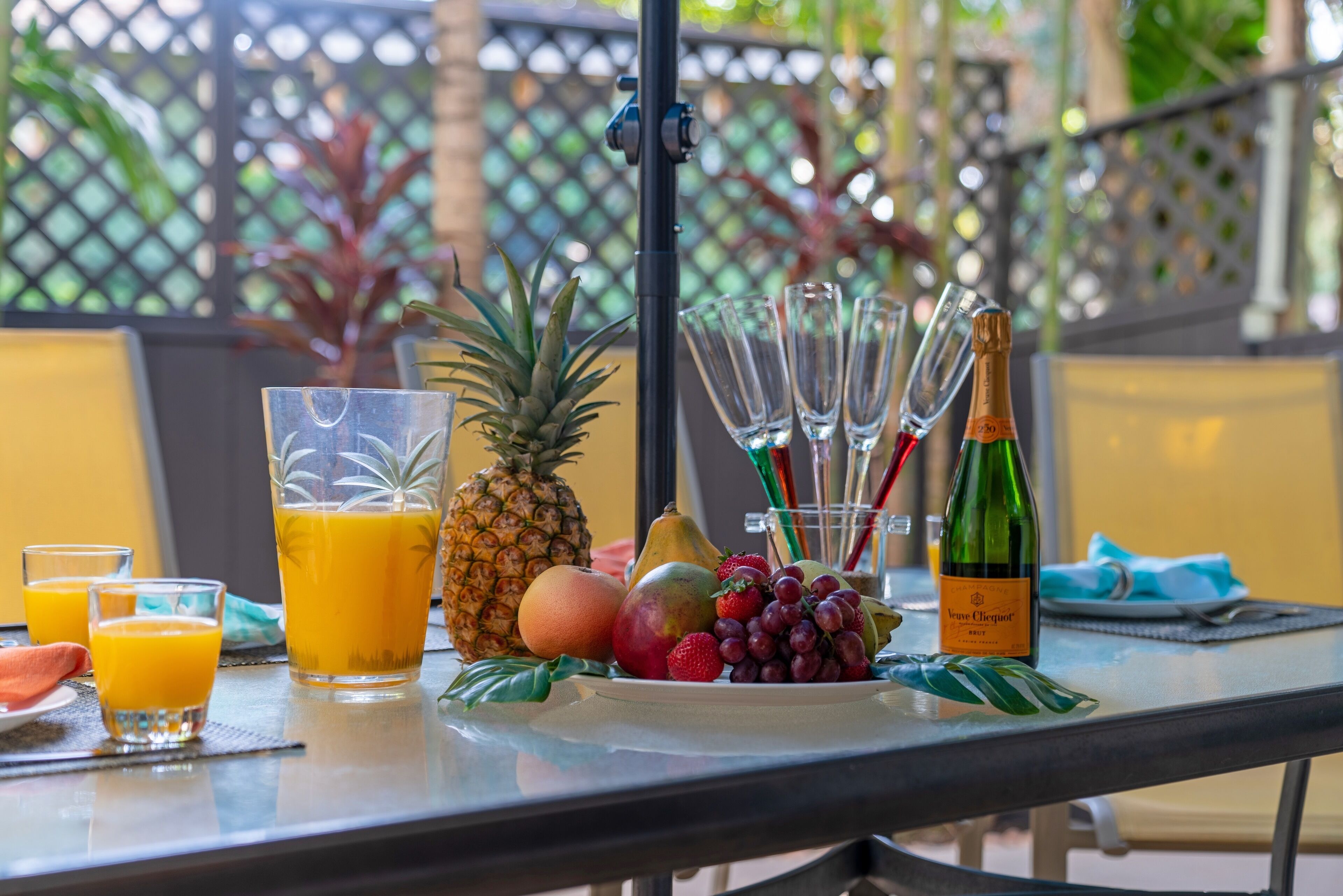 Une table avec un verre de jus d’orange, un ananas, une assiette de fruits, une bouteille de champagne et des verres vides.