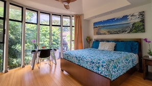 A bedroom with a large bed, a desk with a chair, a ceiling fan, and large windows with a view of greenery.