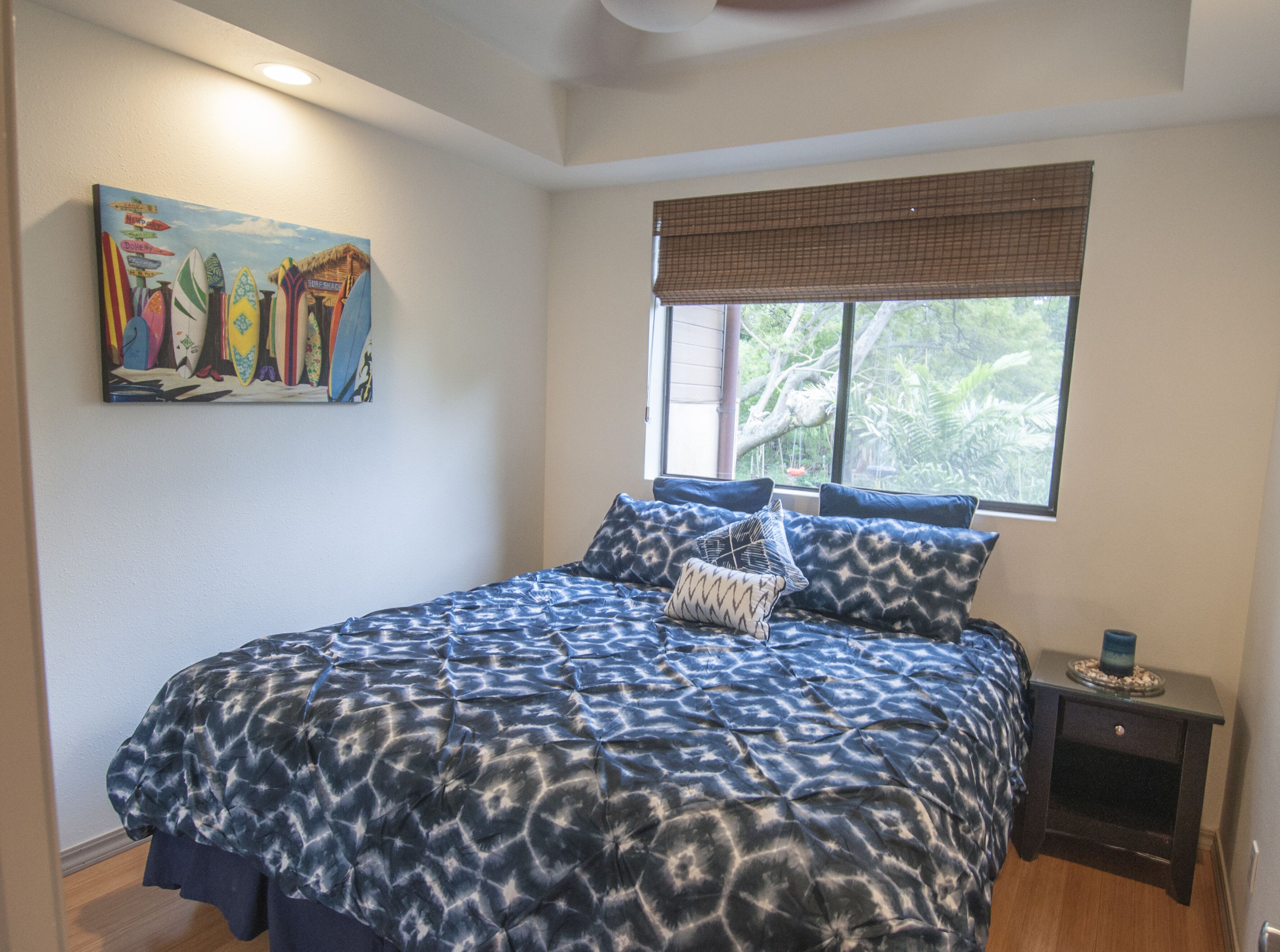 A bedroom with a bed, a window with a view of greenery, a painting on the wall, and a nightstand with a lamp.