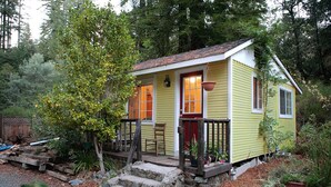 A small yellow house with a red door, a porch, and steps leading up to it. - Raymond's Bakery - Creekside Cabin and Breakfast (Cazadero)