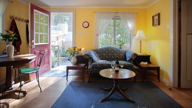 A cozy living room with a sofa, a round coffee table, a dining table with chairs, a lamp, and a clock on the wall. - Raymond's Bakery - Creekside Cabin and Breakfast (Cazadero)
