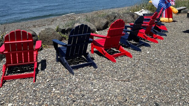 A row of red and black Adirondack chairs on a pebbled beach with a view of the ocean and a forested hillside.