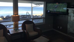 A living room with a large window overlooking the beach, a flat-screen TV mounted on the wall, and a small table with a lamp.