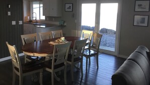 A dining area with a wooden table and white chairs, a ceiling fan, and a view of the beach through large windows.