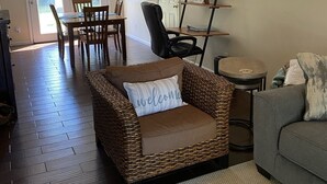 Un salon comprenant un fauteuil en osier, un canapé gris, une table à manger en bois avec des chaises, une bibliothèque et une porte donnant sur l’extérieur.
