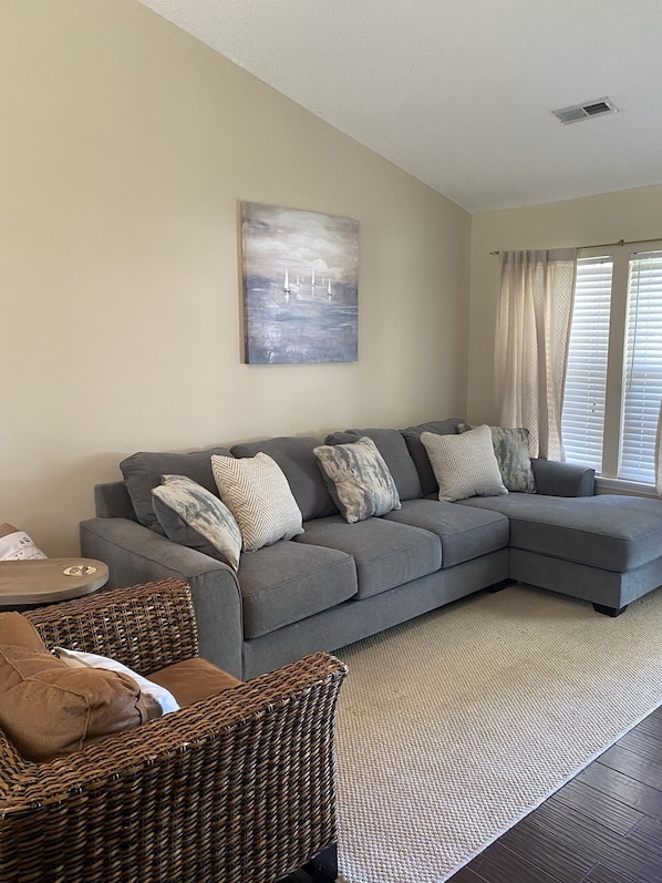 A living room with a grey sectional sofa, a wicker chair, a round coffee table, and a painting of sailboats on the wall.