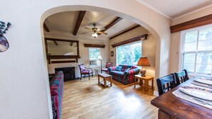 A spacious living area with a fireplace, a sofa, a dining table, and a window with a view of trees. - Great Location in Cheyenne Cañyon, 3Bd, 1.5Bath, Private fenced yard and deck! (Colorado Springs)