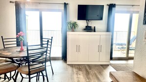 A dining area with a round table, chairs, a vase with flowers, a ceiling fan, a flat-screen TV mounted on the wall, and a sliding glass door leading to an outdoor balcony. - Beachfront View from 9th Floor (South Padre Island)