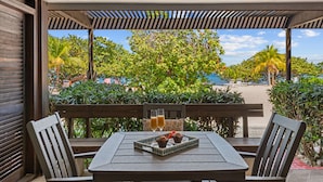 A covered outdoor seating area with a table and chairs, overlooking a beach with palm trees and lounge chairs. - Sapphire Beach, Beach Level Condo, B Bldg, Bella Mar, Remodeled 2025, Generator (St. Thomas)